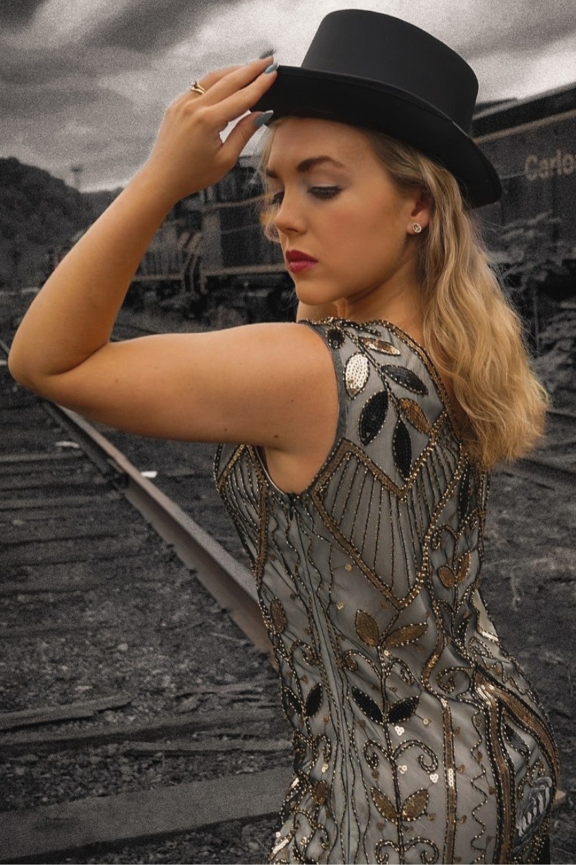 A white woman with blonde hair, a patterned dress, and a fedora hat posing with her hand on her head in front of train tracks