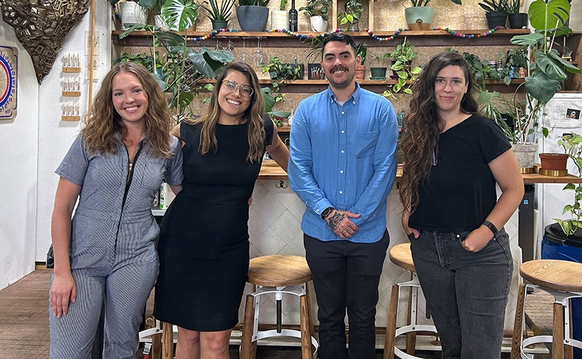 Four white people -- three women and one man -- stand in a row side-by-side smiling for the camera inside a space filled with artwork and plants. 