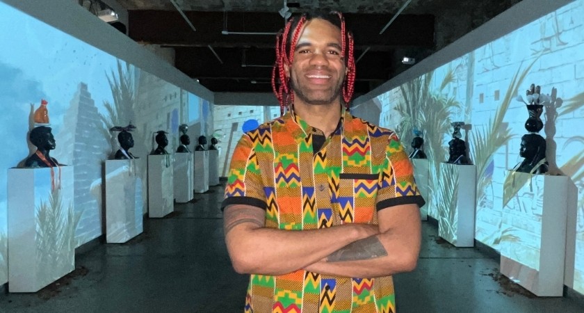 Smiling Black man with red-and-black shoulder-length braids and wearing a colorful African-inspired patterned shirt, poses for the camera while crossing his arms in front of an art gallery exhibit